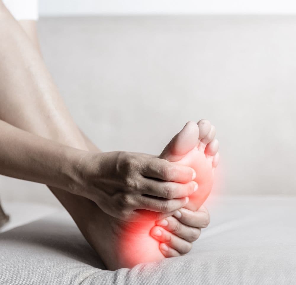 Dłonie masują podeszwę stopy, na której zaznaczono czerwone punkty bólowe w okolicach pięty i śródstopia.