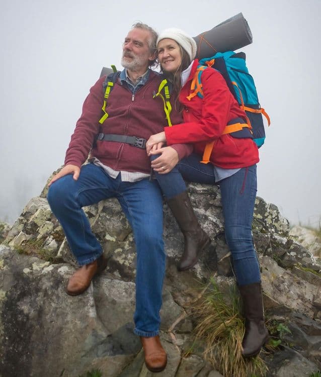 Starsza para w stroju turystycznym, z plecakami, siedząca na skalistym wzniesieniu w mglisty dzień, spoglądająca w dal z uśmiechem i czułością.