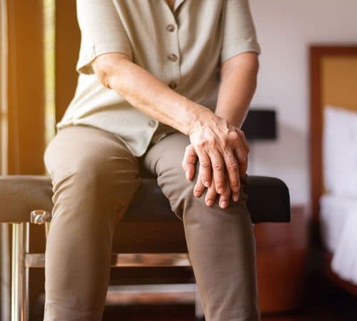 Osoba starsza siedzi na kozetce i obiema rękami trzyma się za kolano w geście odczuwanego dyskomfortu.