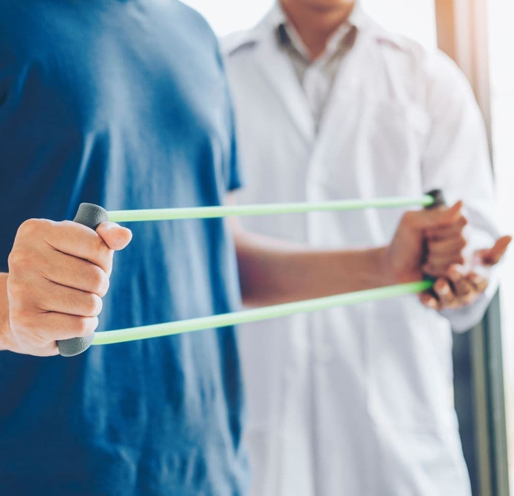 Pacjent trzyma zieloną gumę do ćwiczeń pod nadzorem terapeuty w białym fartuchu. Ćwiczenie ma na celu wzmocnienie siły mięśniowej kończyn górnych.