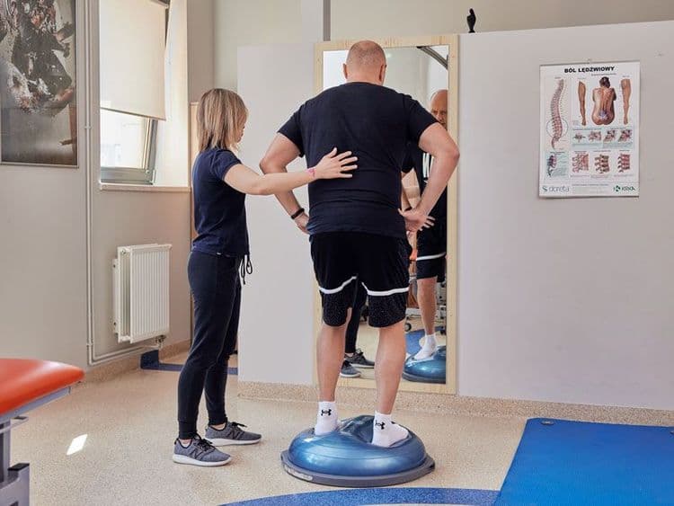 Fizjoterapeutka pomaga pacjentowi utrzymać równowagę podczas ćwiczeń na niebieskiej półkuli Bosu przed lustrem w gabinecie kinezyterapii.