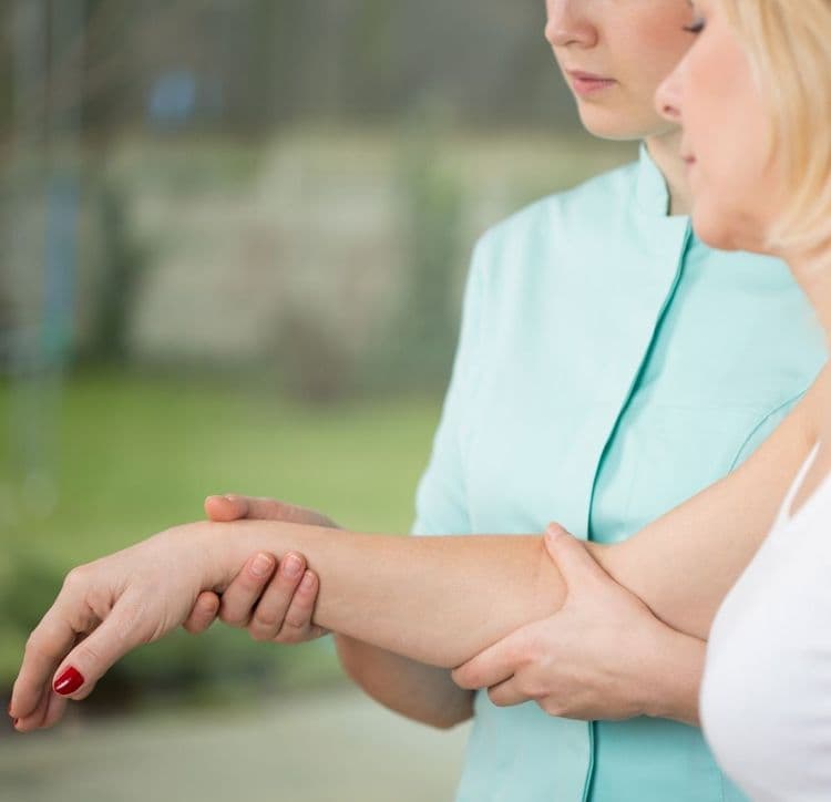 Fizjoterapeutka w miętowym stroju podtrzymuje i bada przedramię oraz nadgarstek pacjentki, sprawdzając zakres ruchów i reakcje neurologiczne.