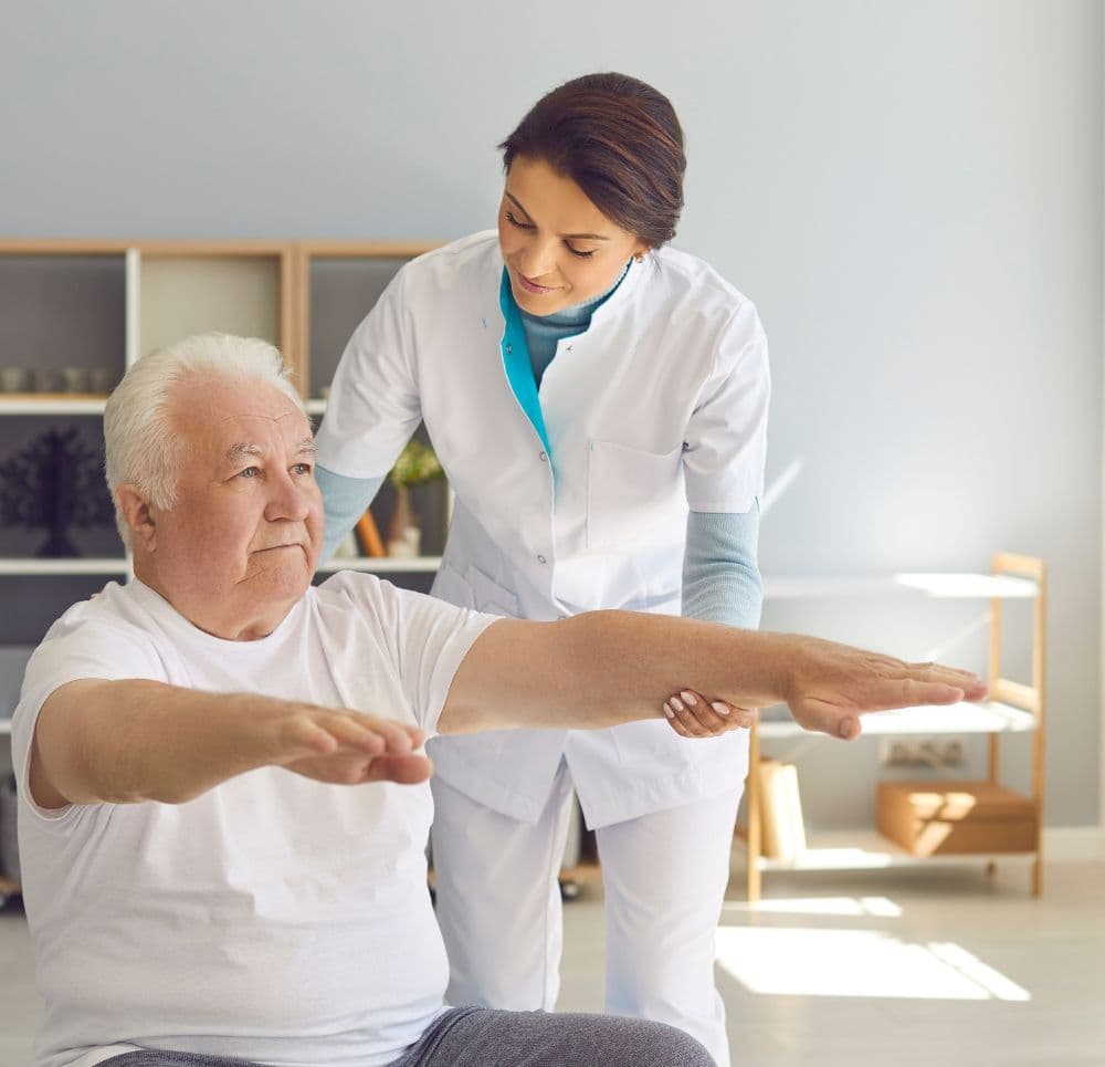 Terapeutyka asystuje starszemu mężczyźnie, który siedząc, wyciąga ręce przed siebie w celu trenowania stabilizacji tułowia i kontroli motorycznej.