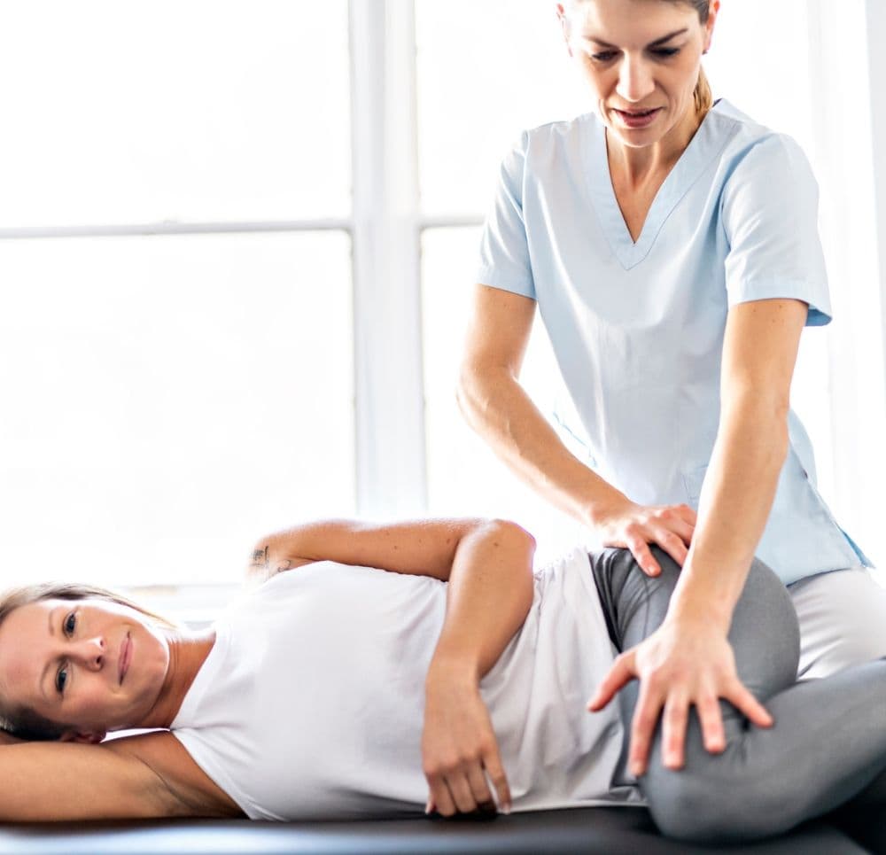 Specjalistka wykonuje terapię manualną na biodrze pacjentki leżącej na boku, pomagając w torowaniu prawidłowych wzorców ruchowych nogi.