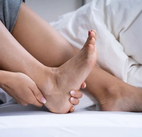 kobieta trzyma w dłoniach swoją stopę, siedząc na łóżku