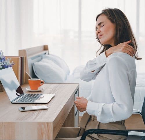 obieta siedząca przy biurku z laptopem, trzymająca się za bolący bark, co obrazuje objawy rwy barkowej spowodowane uciskiem nerwów.